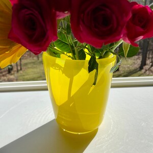 May include: A yellow glass vase with a bouquet of red roses and a yellow flower. The vase is sitting on a white surface.