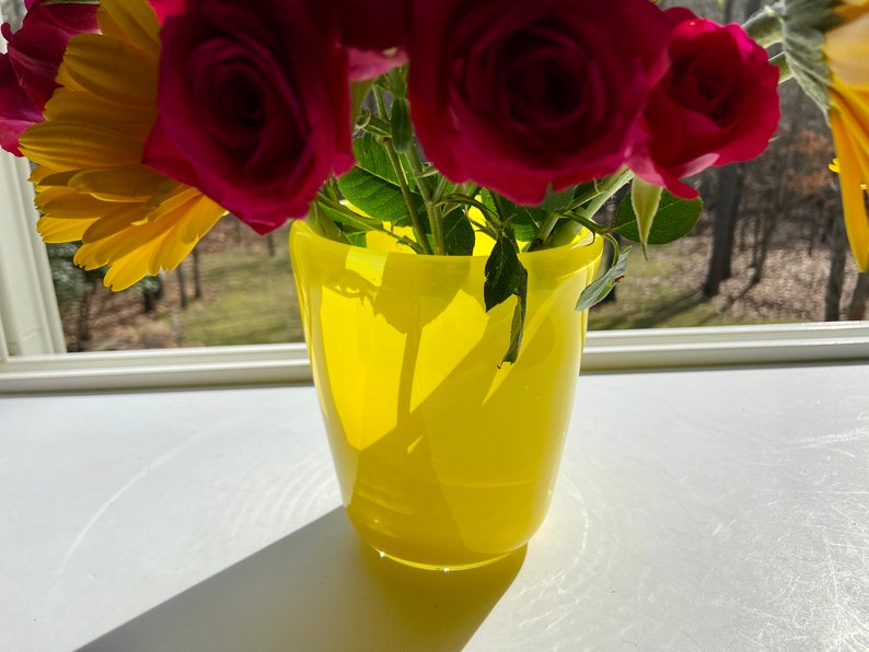 May include: A yellow glass vase with a bouquet of red roses and a yellow flower. The vase is sitting on a white surface.