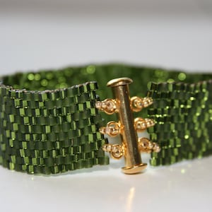 May include: A wide bracelet crafted from small, olive green beads. The beads are woven together in a rectangular pattern. The bracelet features a gold-colored clasp with a cylindrical bar and decorative accents.