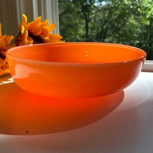 May include: An orange glass bowl with a rounded shape. The bowl is sitting on a white surface, with a shadow cast by the sunlight. In the background, there are yellow sunflowers and a window with a view of green trees.