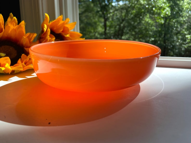May include: An orange glass bowl with a rounded shape. The bowl is sitting on a white surface, with a shadow cast by the sunlight. In the background, there are yellow sunflowers and a window with a view of green trees.