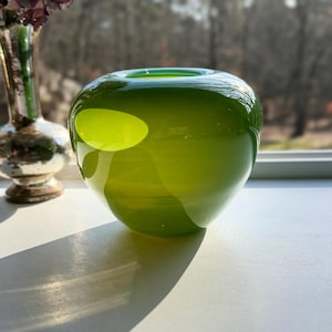 Puede incluir: Un jarrón redondo de vidrio verde con una abertura ancha. El jarrón tiene un acabado liso y brillante y una forma ovalada verde claro en el lateral. Un jarrón plateado y flores secas están en el fondo.