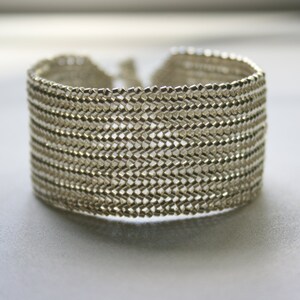 May include: A silver bracelet made of small, interconnected metal links, creating a woven, textured pattern.