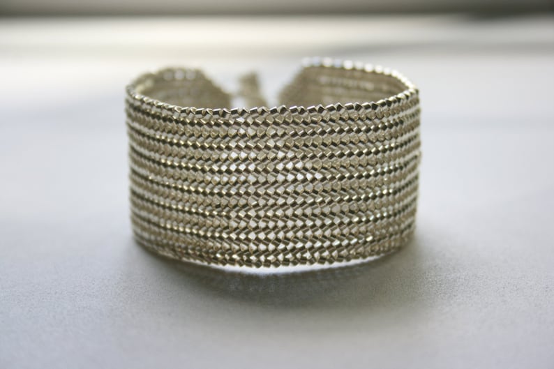 May include: A silver bracelet made of small, interconnected metal links, creating a woven, textured pattern.