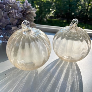 May include: Two white glass pumpkins with a textured surface. The pumpkins are sitting on a white surface.