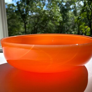 May include: An orange glass bowl with a rounded shape. The bowl is sitting on a white surface, with a blurred background of green trees and a window. The bowl is made of a translucent material.