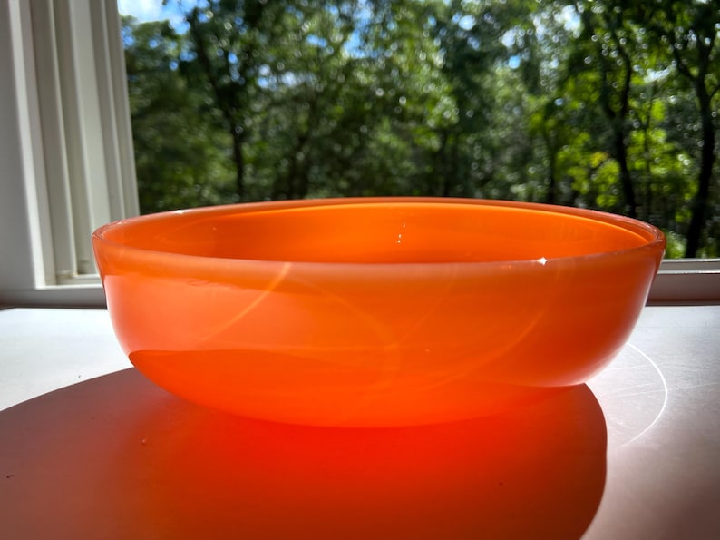 May include: An orange glass bowl with a rounded shape. The bowl is sitting on a white surface, with a blurred background of green trees and a window. The bowl is made of a translucent material.