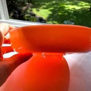 May include: An orange glass bowl with a rounded shape. The bowl is held by a hand near a window. The bowl is reflecting the light and the color is vibrant. The bowl is a decorative item.