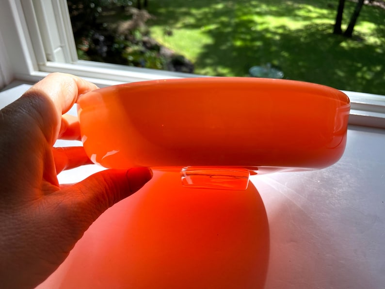 May include: An orange glass bowl with a rounded shape. The bowl is held by a hand near a window. The bowl is reflecting the light and the color is vibrant. The bowl is a decorative item.