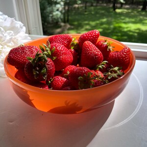 May include: A vibrant orange bowl filled with fresh, ripe strawberries. The bright red berries have green leafy tops and are clustered together. A large white flower is visible in the background, near a window with a view of green trees.