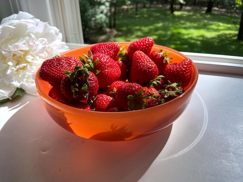 May include: A vibrant orange bowl filled with fresh, ripe strawberries. The bright red berries have green leafy tops and are clustered together. A large white flower is visible in the background, near a window with a view of green trees.