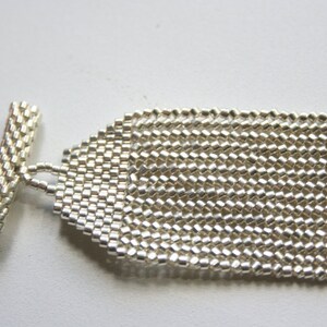 May include: A silver beaded bracelet with a toggle clasp. The bracelet is made of small, square beads that are woven together in a pattern.