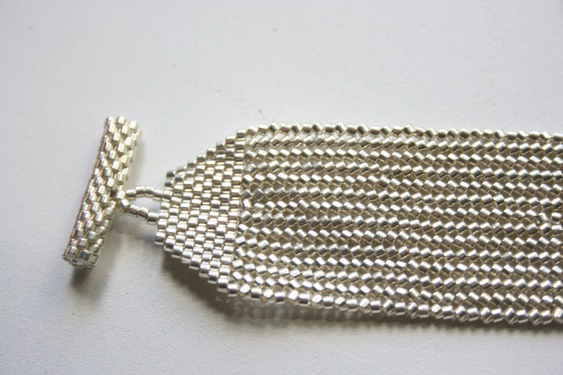 May include: A silver beaded bracelet with a toggle clasp. The bracelet is made of small, square beads that are woven together in a pattern.