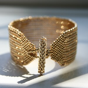 May include: A gold-colored bracelet made of small, interconnected metal beads. The bracelet has a unique clasp design that resembles a bar.