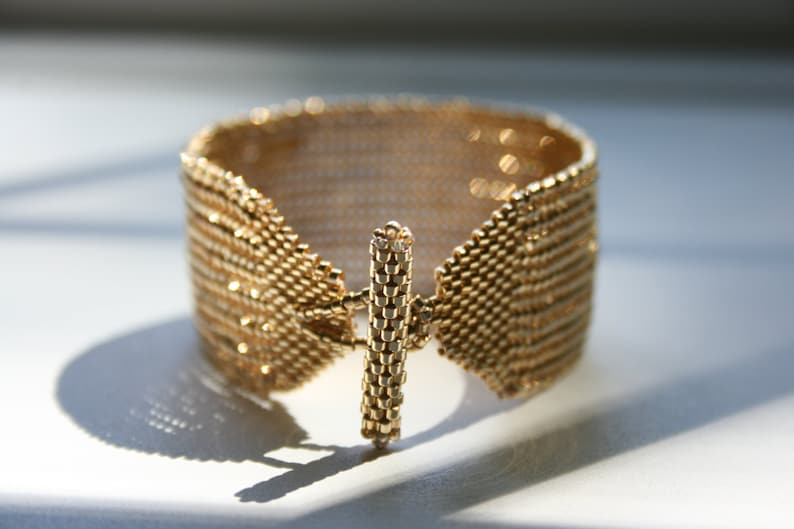 May include: A gold-colored bracelet made of small, interconnected metal beads. The bracelet has a unique clasp design that resembles a bar.