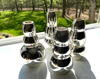 Puede incluir: Cuatro candelabros de vidrio plateado y espejo de diferentes tamaños y formas. Los candelabros están dispuestos sobre una superficie blanca.