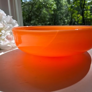 May include: An orange glass bowl with a rounded shape. The bowl is sitting on a white surface, with a large white flower and green foliage in the background. The bowl's shadow is visible on the surface.