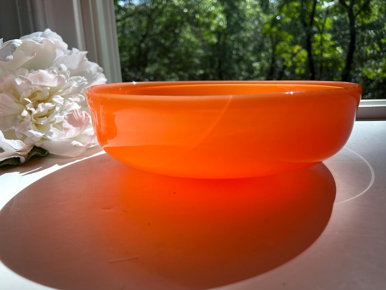 May include: An orange glass bowl with a rounded shape. The bowl is sitting on a white surface, with a large white flower and green foliage in the background. The bowl's shadow is visible on the surface.