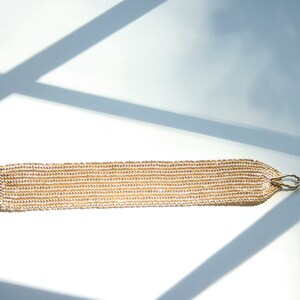 May include: A gold beaded bracelet with a toggle clasp. The bracelet is made of small, round beads that are strung together in a chain-like pattern.