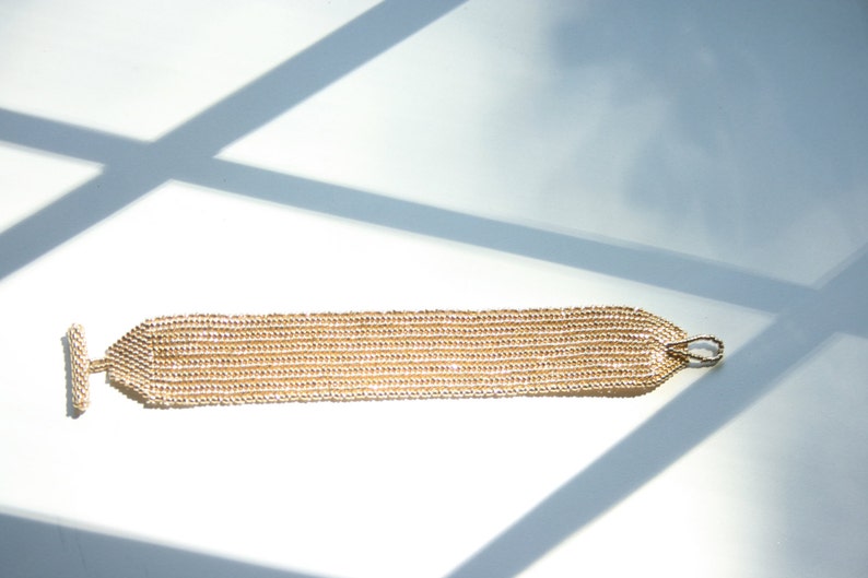May include: A gold beaded bracelet with a toggle clasp. The bracelet is made of small, round beads that are strung together in a chain-like pattern.