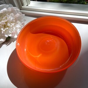 May include: An orange glass bowl with a rounded shape. The bowl is sitting on a white surface next to a large white flower. The bowl's shadow is visible on the surface.