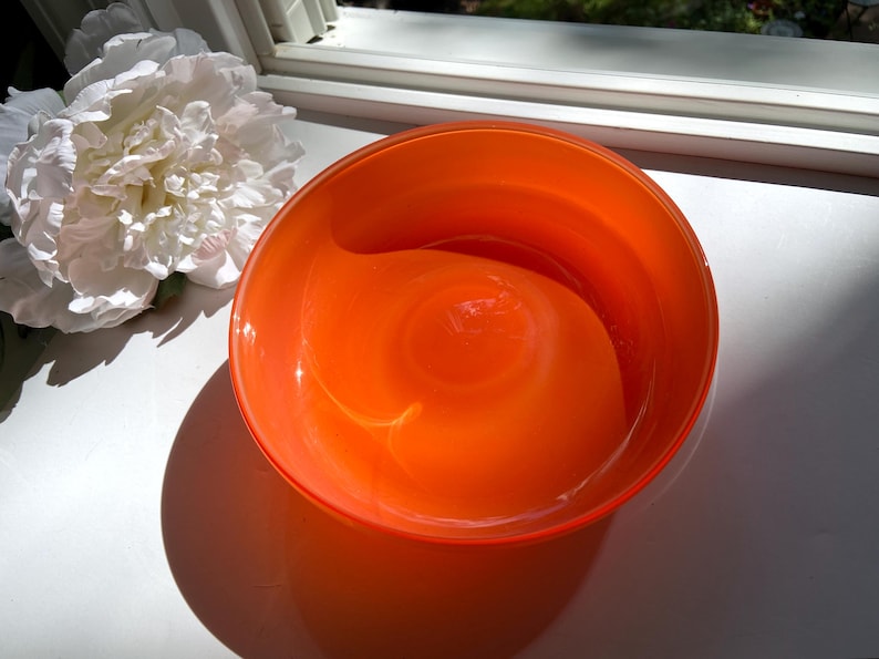 May include: An orange glass bowl with a rounded shape. The bowl is sitting on a white surface next to a large white flower. The bowl's shadow is visible on the surface.