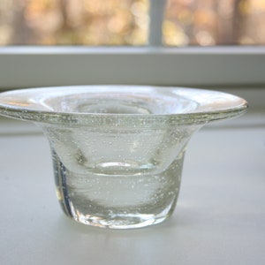 May include: A clear glass candle holder with a wide, flat top and a round, flared base. The glass has a textured surface with small air bubbles.