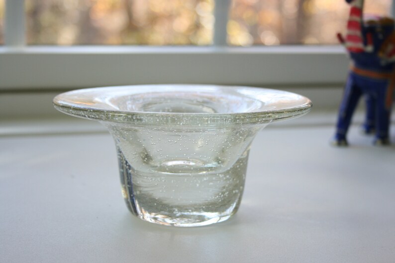 May include: A clear glass candle holder with a wide, flat top and a round, flared base. The glass has a textured surface with small air bubbles.