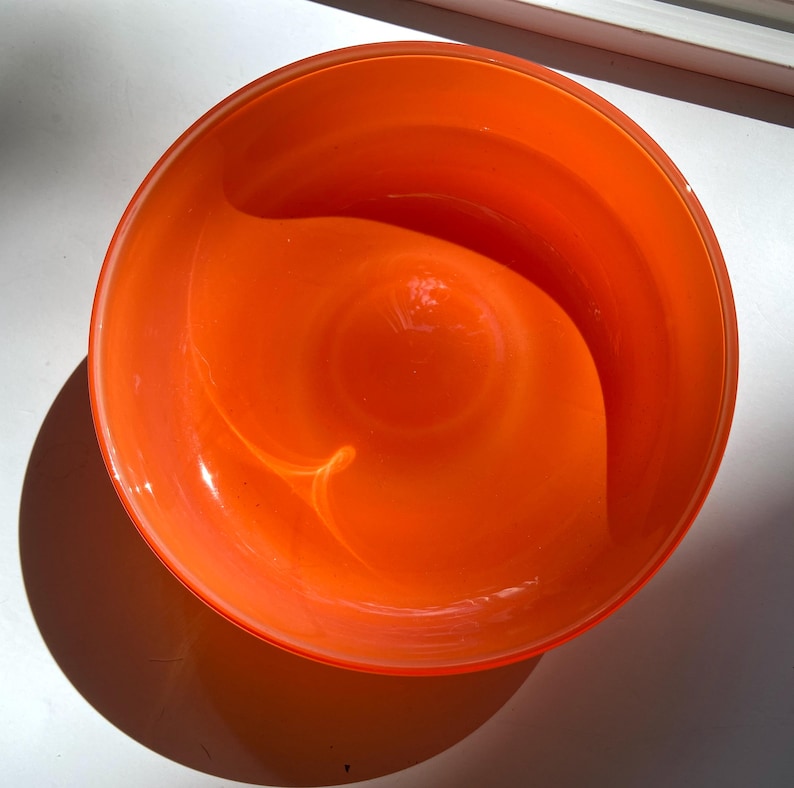 May include: A vibrant orange glass bowl with a rounded shape. The bowl has a glossy finish and a smooth interior. The light creates a swirl pattern in the center. The bowl is likely used for decorative purposes or serving.