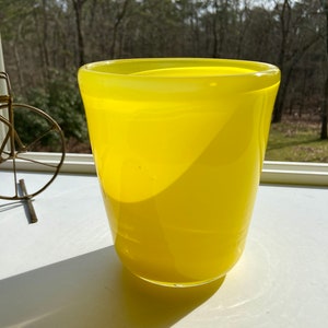 May include: A bright yellow glass vase with a rounded bottom and a wide opening. The vase is sitting on a white surface.