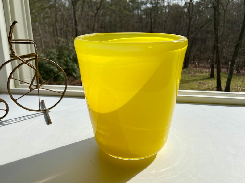 May include: A bright yellow glass vase with a rounded bottom and a wide opening. The vase is sitting on a white surface.