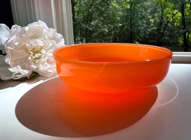 May include: An orange glass bowl with a rounded shape. The bowl is sitting on a white surface, with a white flower and green foliage in the background. The bowl casts a large shadow.