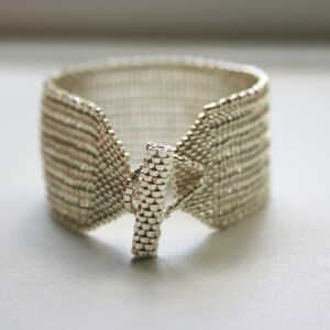 May include: A silver beaded bracelet with a toggle clasp. The bracelet is made of small, round beads that are woven together in a pattern. The clasp is a simple, rectangular piece of metal.