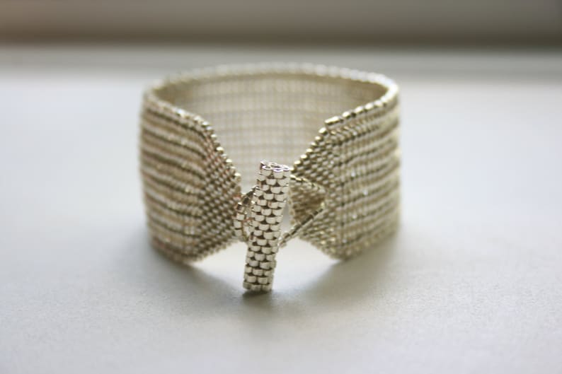May include: A silver beaded bracelet with a toggle clasp. The bracelet is made of small, round beads that are woven together in a pattern. The clasp is a simple, rectangular piece of metal.
