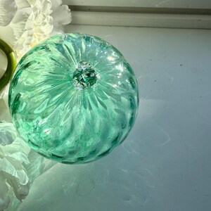 Op de afbeelding: Een rond, lichtgroen decoratief object van glas met een textuur. Het object heeft een kleine, heldere glazen knop aan de bovenkant en bevindt zich in de buurt van witte bloemen.
