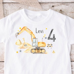 Könnte beinhalten: Weißes T-Shirt mit einem gelben Bagger, einer Schaufel und einem Baustellenkegel. Das Shirt trägt außerdem den Namen "Levi" und die Zahl "4" aufgedruckt.