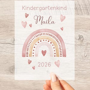 Könnte beinhalten: Ein rechteckiger Druck mit dem Text Kindergartenkind, dem Namen Maila und dem Jahr 2026. Das Design zeigt einen Regenbogen im Aquarellstil in Rosa- und Beigetönen, verziert mit kleinen Herzen und gepunkteten Linien auf weißem Hintergrund.