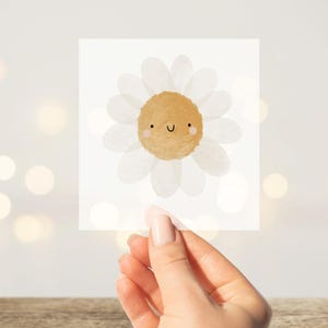 Könnte beinhalten: Eine quadratische Karte mit einer Aquarellillustration einer lächelnden Gänseblümchen. Das Gänseblümchen hat weiße Blütenblätter und eine hellbraune Mitte. Die Karte wird von einer Hand gehalten, mit einem Holztisch im Vordergrund und einem verschwommenen Hintergrund mit Bokeh-Lichtern.