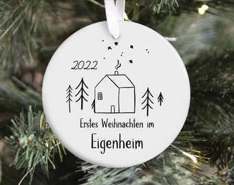 May include: White ceramic Christmas ornament with a black and white house illustration, trees, stars, and the year 2022. The ornament has a white ribbon for hanging and is displayed on a Christmas tree.
