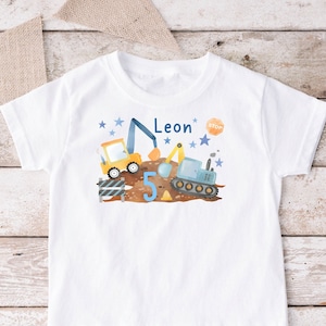 Könnte beinhalten: Weißes T-Shirt mit einem blauen und gelben Baufahrzeug-Design. Das Design beinhaltet einen Bagger, einen LKW und ein Stoppschild. Die Zahl 5 ist ebenfalls im Design enthalten. Der Text "Leon" ist auf dem Shirt gedruckt.