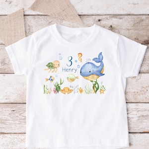 Puede incluir: Camiseta blanca con una ballena azul, tortugas marinas, peces y otras criaturas marinas. El número 3 y el nombre Henry están impresos en la camiseta.