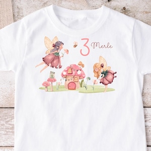 Könnte beinhalten: Weißes T-Shirt mit einer rosa und grünen Feen-Illustration, einem Pilzhaus und dem Text "3 Merle".