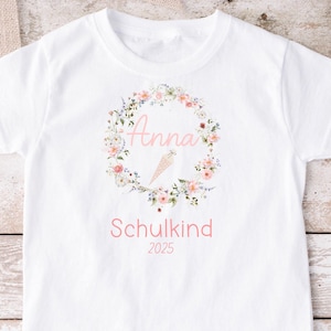Puede incluir: Camiseta blanca con un diseño de corona de flores y el nombre "Anna" en letras rosas. El texto "Schulkind 2025" también está impreso en la camiseta.