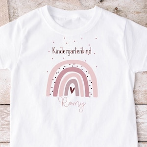 Può includere: Maglietta bianca con un design arcobaleno rosa e marrone e il testo "Kindergartenkind" e "Romy".