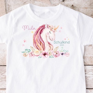 Könnte beinhalten: Weißes T-Shirt mit einer pinkfarbenen und gelben Einhorn-Illustration, Blumen und Sternen. Der Text "Mila" und "Schulkind 2023" ist auf dem Shirt gedruckt.