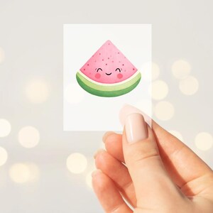 Könnte beinhalten: Ein klarer, rechteckiger Aufkleber mit einem lächelnden Wassermelonenstück. Die Wassermelone hat ein rosa Inneres, schwarze Samen und eine grüne Schale. Der Aufkleber wird von einer Hand vor einem verschwommenen Hintergrund gehalten.