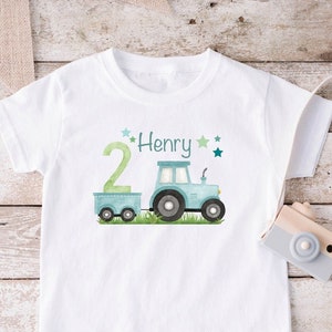 Pode incluir: Camiseta branca com um trator azul e um reboque, grama verde e o número 2. O texto "Henry" está acima do trator. Há pequenas estrelas verdes ao redor do texto.