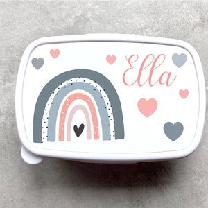 Op de afbeelding: Witte lunchbox met een regenboogontwerp in tinten grijs, roze en wit. De regenboog heeft zwarte stippen. De lunchbox heeft een roze hart in het midden van de regenboog en verschillende roze en grijze harten verspreid over de buitenkant. De naam "Ella" staat in roze cursief op de lunchbox.