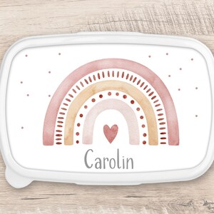 Op de afbeelding: Witte lunchbox met een roze, oranje en bruin aquarel regenboogontwerp. De regenboog heeft een klein roze hart in het midden. De naam "Carolin" staat onder de regenboog gedrukt.