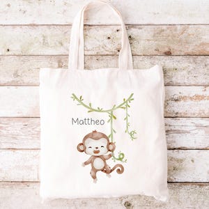 Può includere: Una borsa tote in tela bianca con un'illustrazione di una scimmia marrone appesa a una vite. La scimmia sorride e ha la pelliccia marrone. Il testo "Mattheo" è stampato sulla borsa.